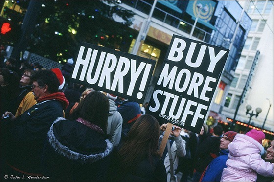 A crowd of people, two of whom are holding placards reading "Hurry!" and "Buy more stuff!".